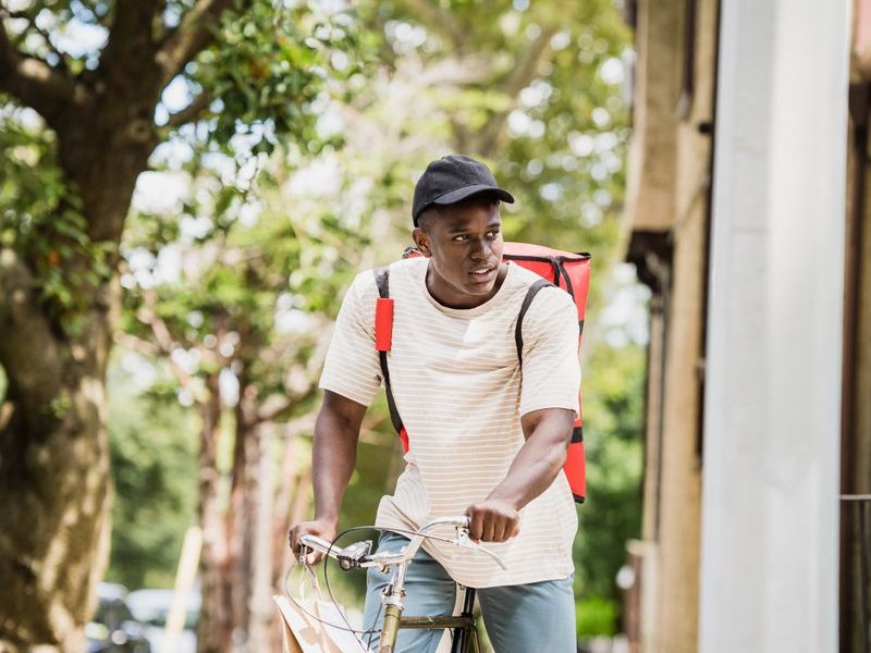 Courier on a bicycle with a red delivery bag riding on a sunlit street
